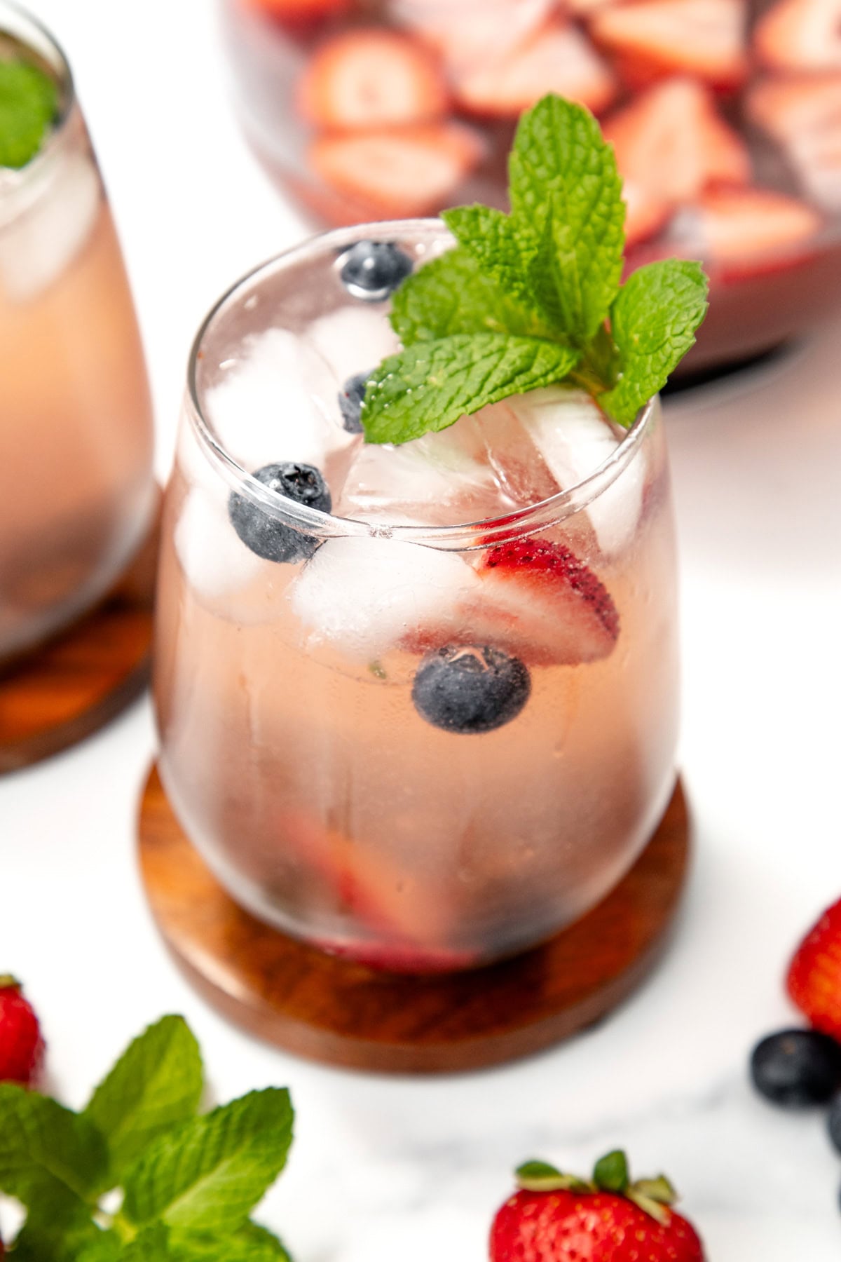 A glass of iced fruity drink with blueberries, strawberries, and mint garnish on a wooden coaster.