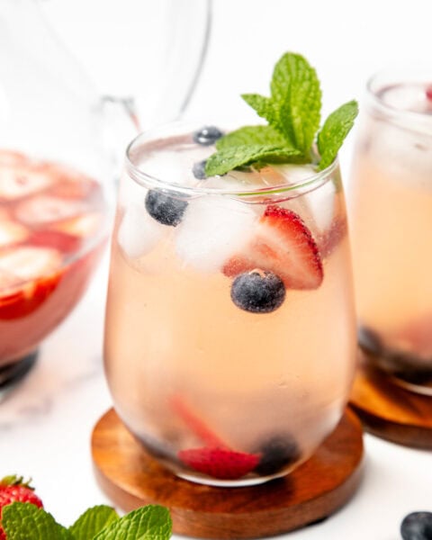 A glass of pink berry-infused drink with ice, blueberries, strawberries, and mint garnish on a wooden coaster.