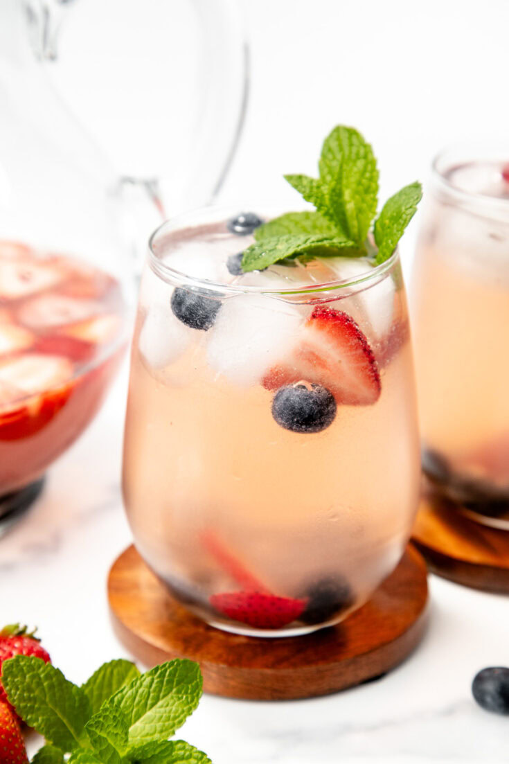 A glass of pink berry-infused drink with ice, blueberries, strawberries, and mint garnish on a wooden coaster.