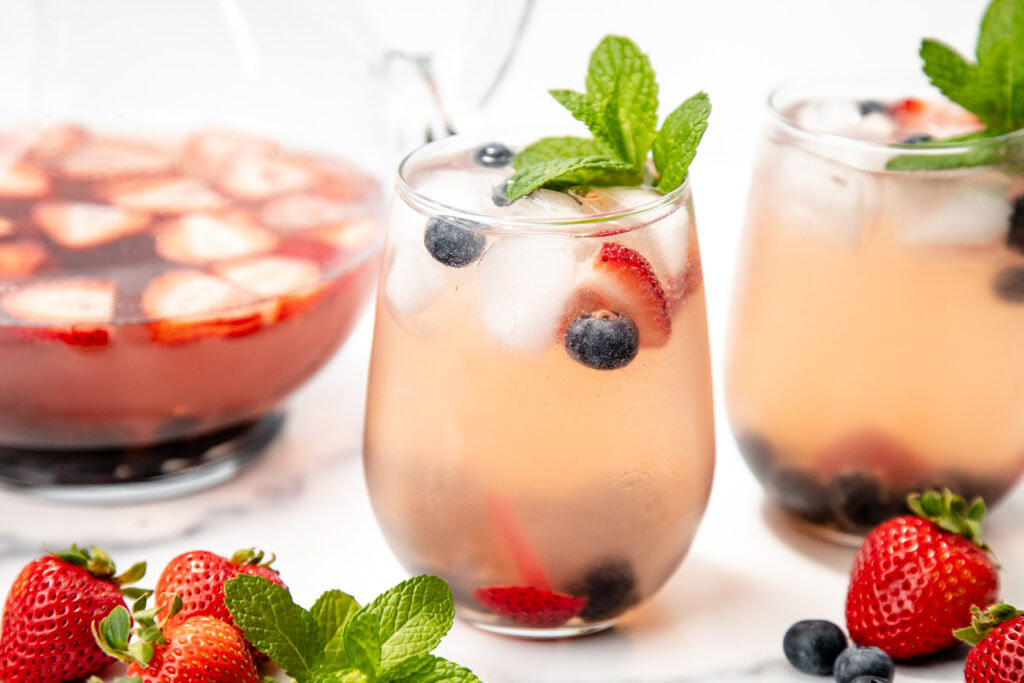Two glasses of berry punch with ice, mint, strawberries, and blueberries; punch bowl and fresh berries in background.
