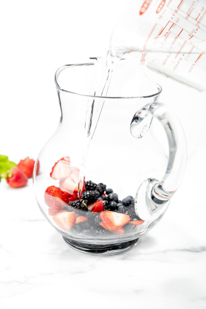 Water being poured from a measuring cup into a glass pitcher filled with strawberries and blackberries.