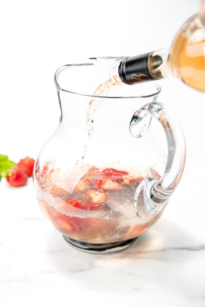 Ros&eacute; wine being poured into a glass pitcher with sliced strawberries on a white surface.