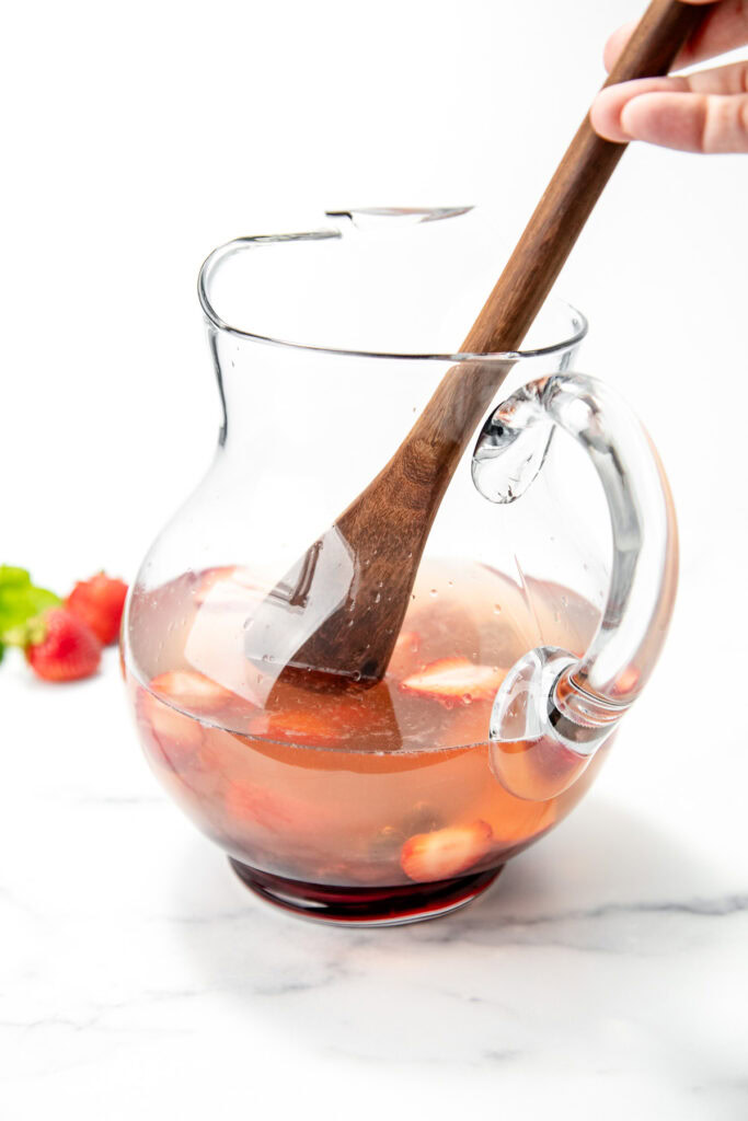 A hand stirs strawberry-infused water in a clear glass pitcher with a wooden spoon.