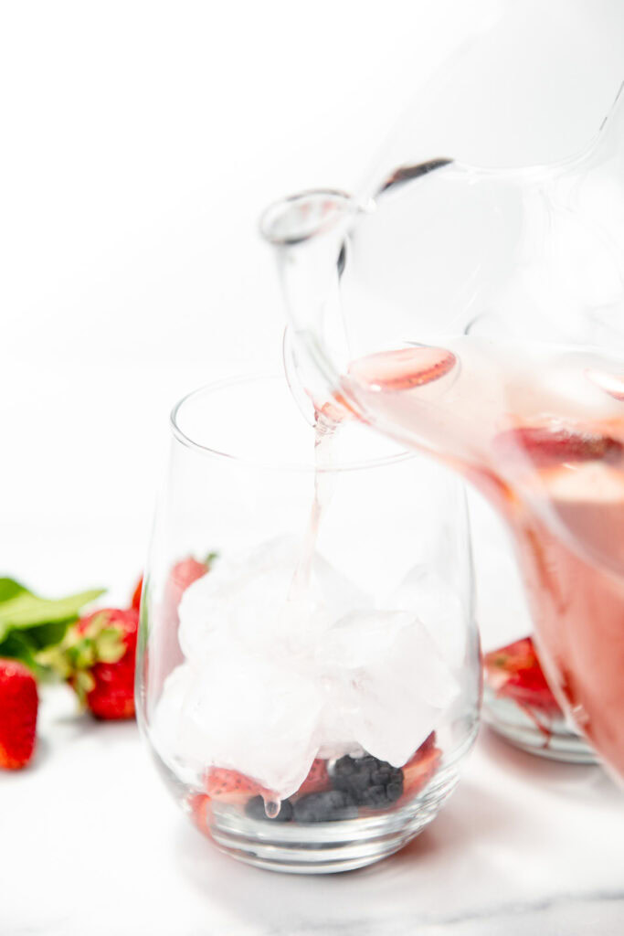 A pitcher pours pink liquid over ice and mixed berries in a glass, with strawberries nearby.