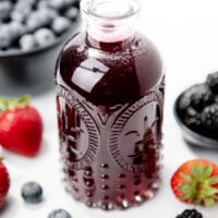 A decorative glass bottle filled with dark berry juice, surrounded by fresh strawberries, blueberries, and blackberries.