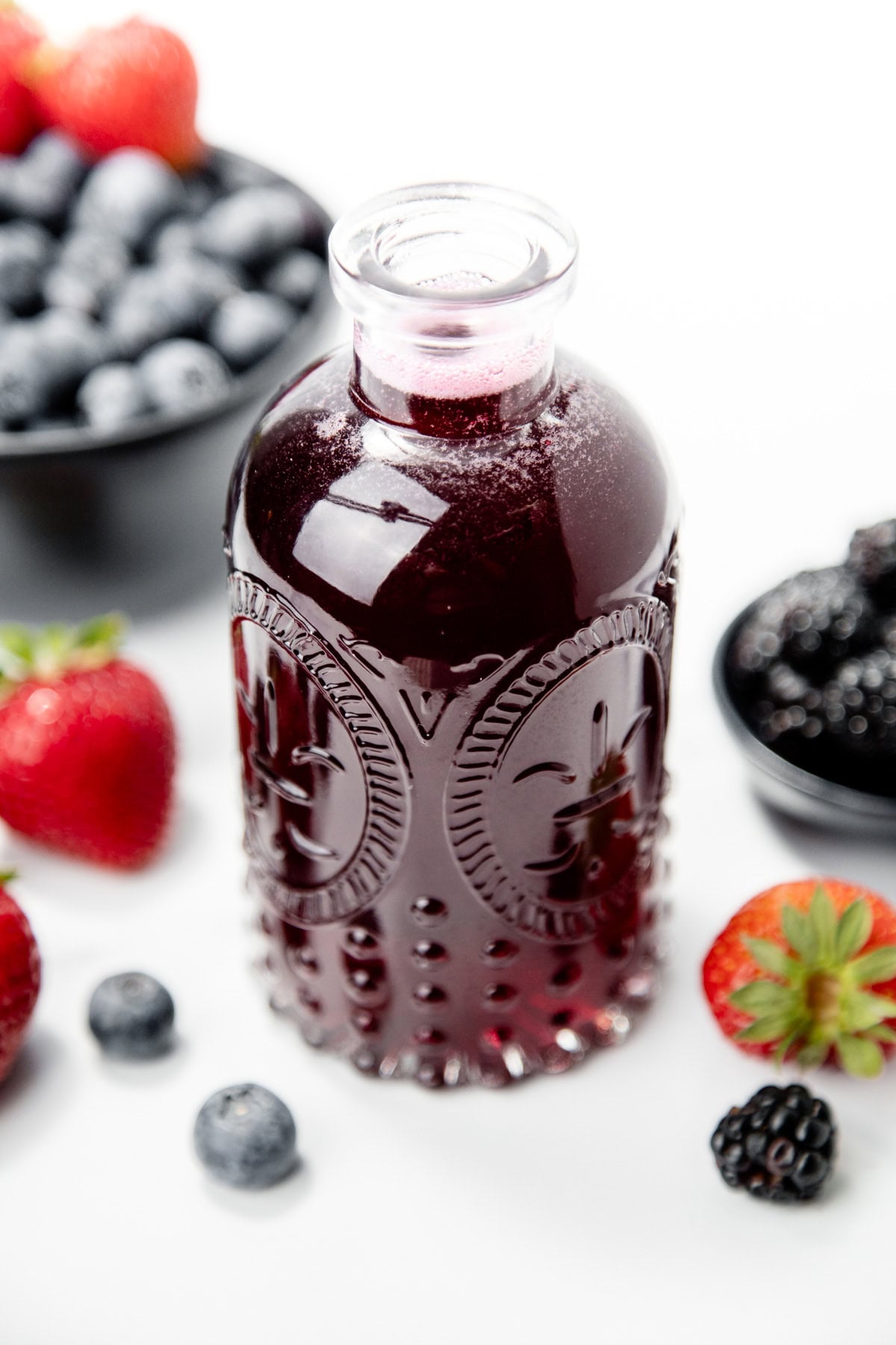A decorative glass bottle filled with dark berry juice, surrounded by fresh strawberries, blueberries, and blackberries.