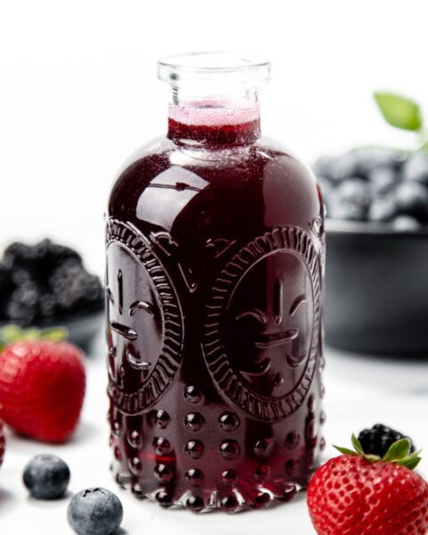 A glass bottle of dark berry juice surrounded by strawberries, blueberries, and blackberries on a white surface.