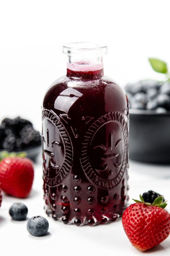 A glass bottle of dark berry juice surrounded by strawberries, blueberries, and blackberries on a white surface.
