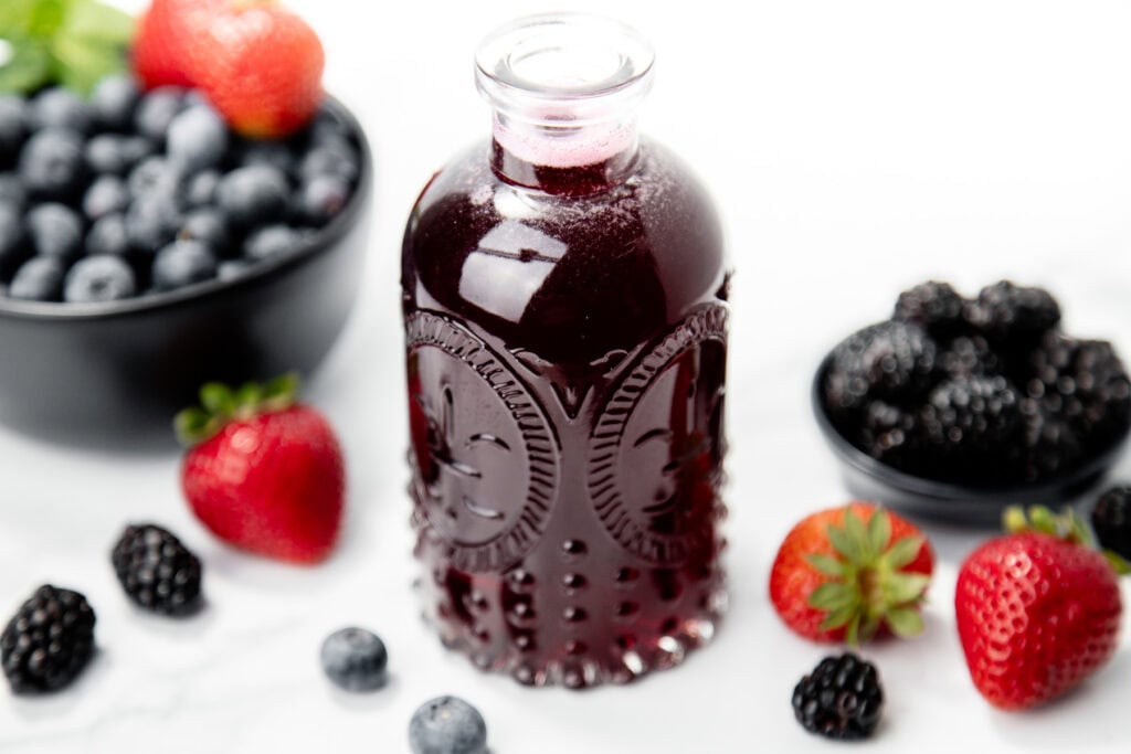 A glass bottle of berry juice surrounded by strawberries, blueberries, and blackberries on a white surface.