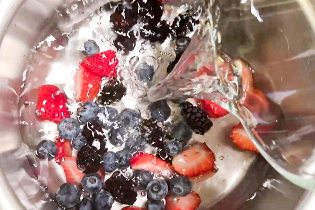 Pouring water over strawberries, blueberries, and blackberries in a stainless steel bowl.