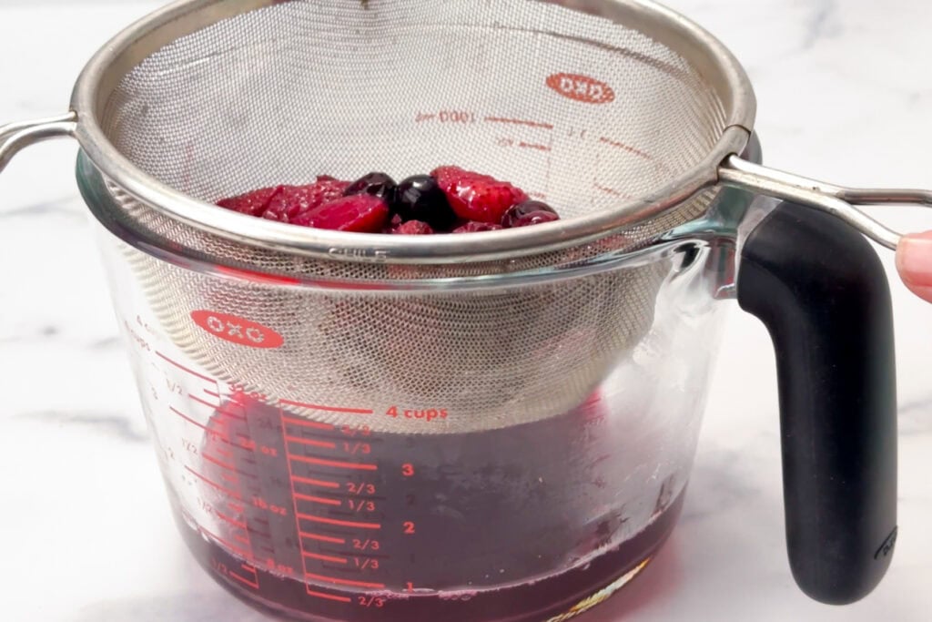 A mesh strainer with berries sits over a glass measuring cup filled with purple juice.