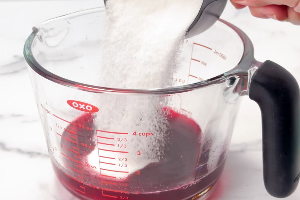 A hand pours sugar into a glass measuring cup filled with red liquid on a white surface.