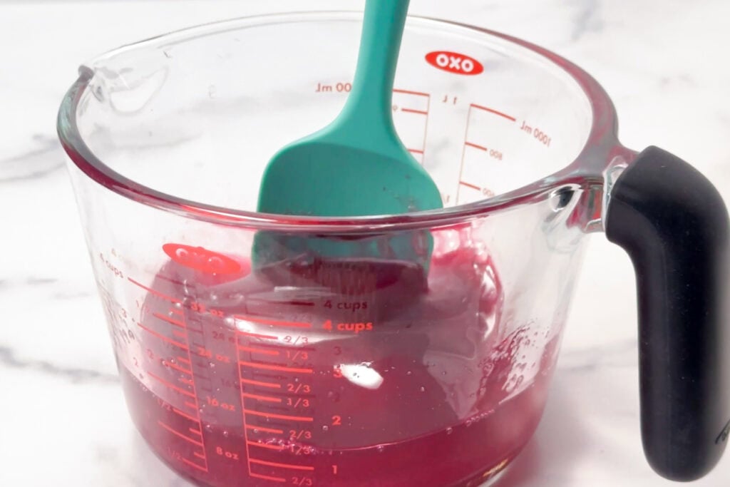 A teal spatula rests in a glass measuring cup with a small amount of red liquid inside.