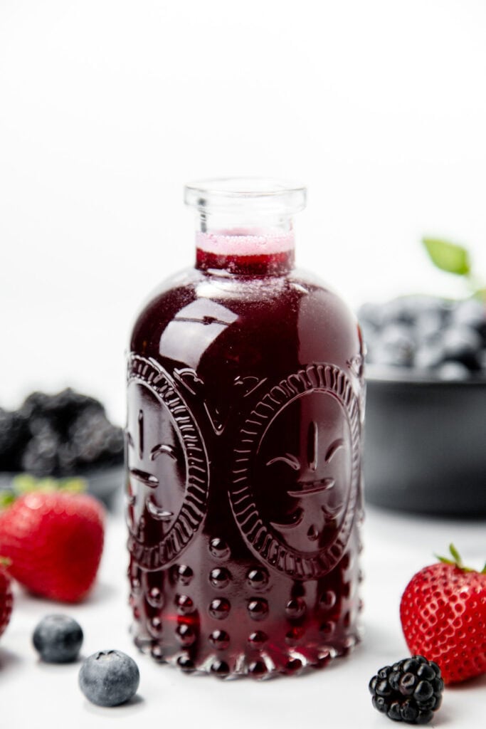 A glass bottle of dark berry juice surrounded by strawberries, blueberries, and blackberries on a white background.