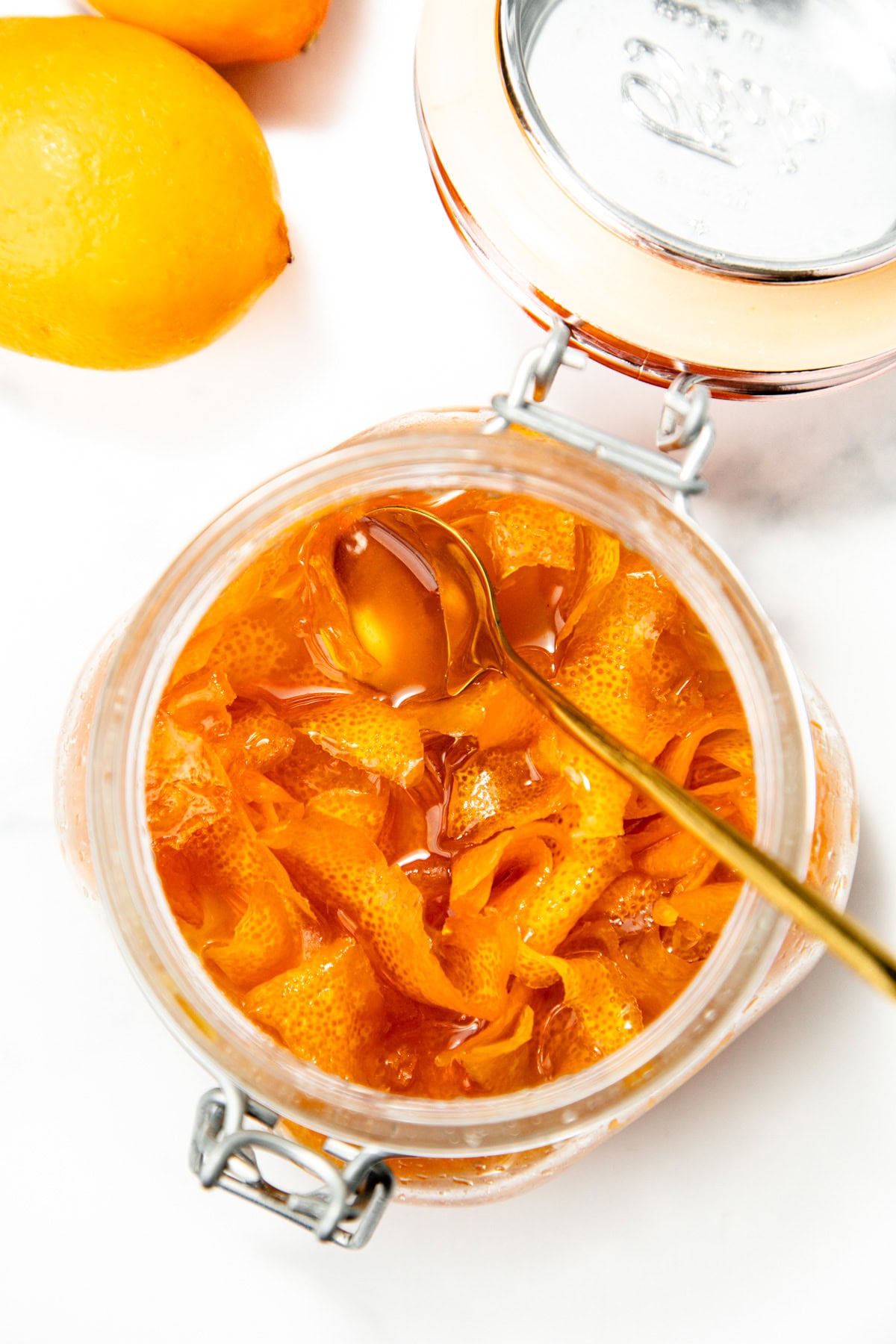 An open glass jar filled with lemon peel marmalade and a spoon, with whole lemons beside it on a white surface.