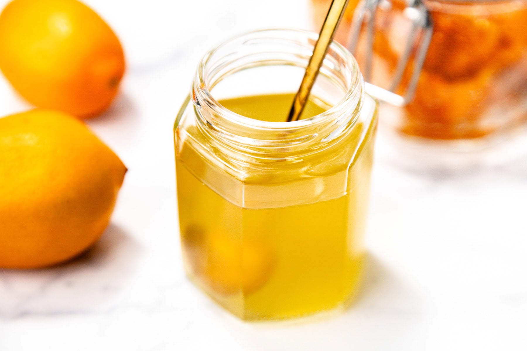 A hexagonal glass jar filled with yellow liquid and a metal spoon, with lemons and another jar in the background on a white surface.