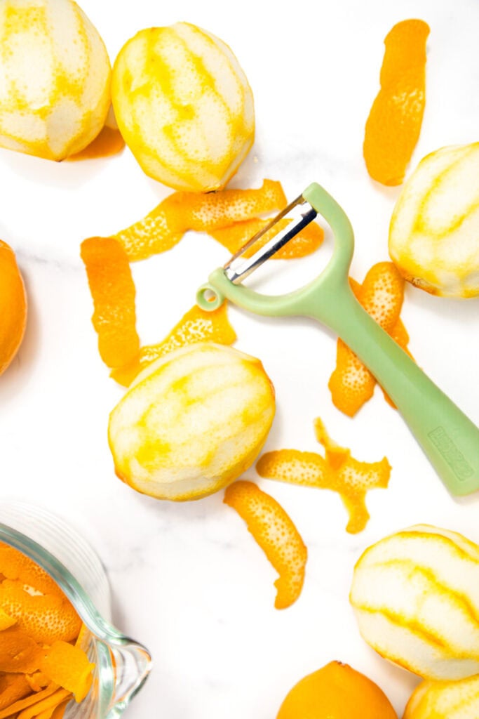 Several peeled lemons, lemon peels, a green vegetable peeler, and a glass bowl with peels are arranged on a white surface.