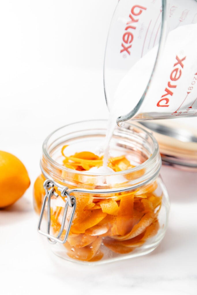 A glass jar filled with orange peels is being sprinkled with granulated sugar from a measuring cup. A whole orange is beside the jar.