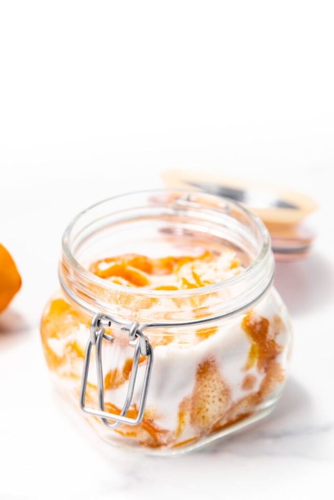 A glass jar filled with orange fruit slices and sugar, with the lid open, placed on a white surface.