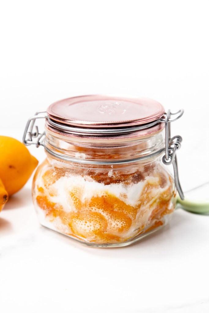 A glass jar with a metal clasp lid contains preserved yellow fruit and sugar, with some foam visible inside. Two pieces of fresh yellow fruit are beside the jar on a white surface.