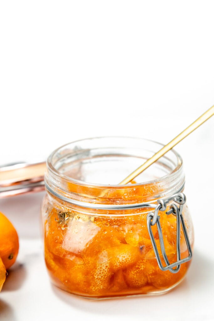 A glass jar filled with orange marmalade, with a spoon inside, sits on a white surface. Some fresh oranges are partially visible on the left.