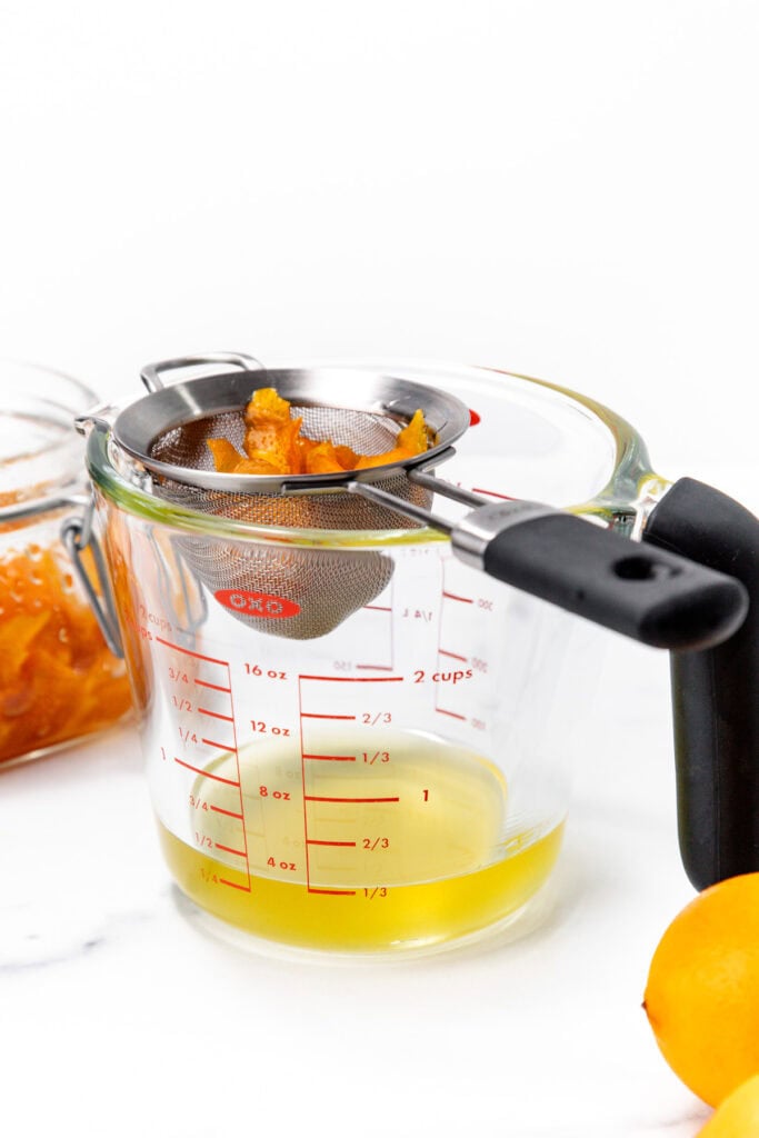A metal strainer with orange peels rests on a glass measuring cup containing yellow liquid, with a jar of peels and a lemon nearby.