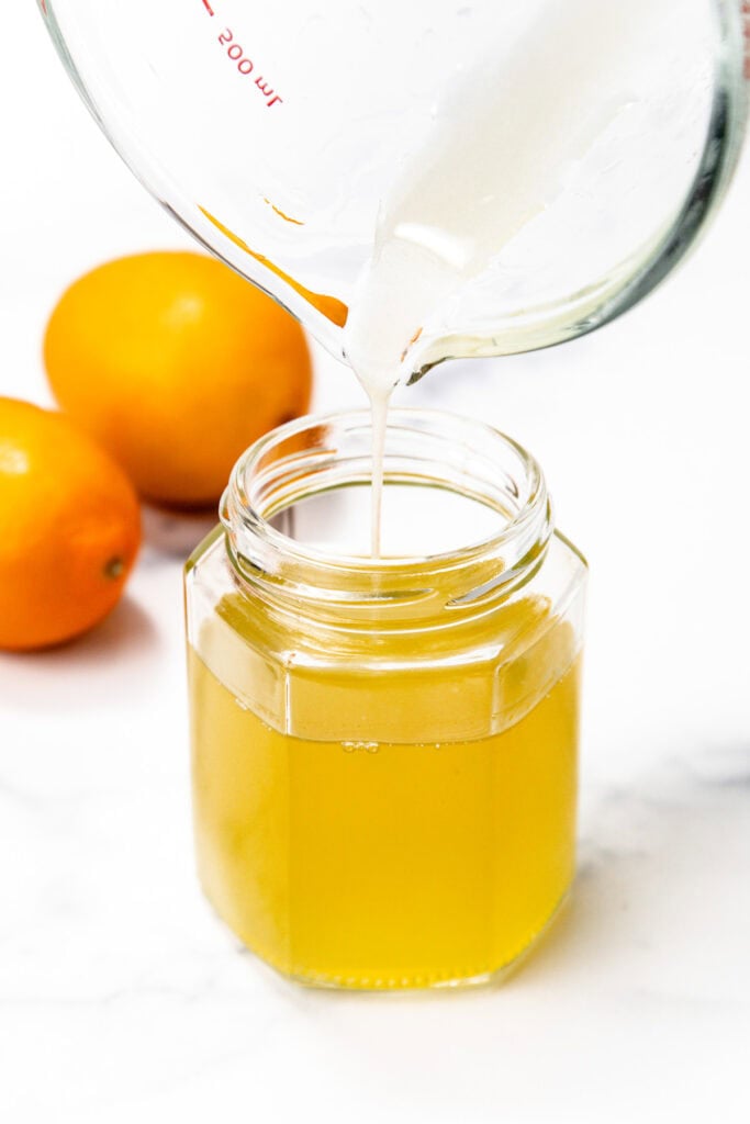 A glass jar filled with yellow liquid, with a white liquid being poured into it from a measuring cup. Two oranges are in the background.