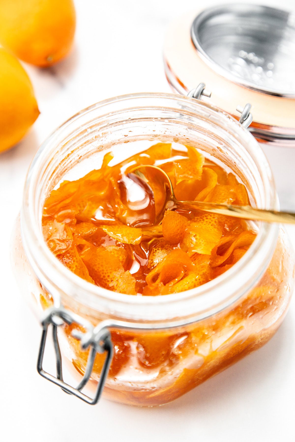 An open glass jar filled with orange marmalade, showing orange peel strips and a spoon inside. Lemons are visible in the background.
