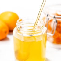 A hexagonal glass jar filled with golden honey, a metal spoon inside, with whole lemons and a jar of preserved lemons in the background.