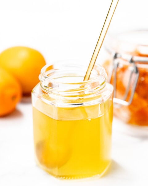A hexagonal glass jar filled with golden honey, a metal spoon inside, with whole lemons and a jar of preserved lemons in the background.