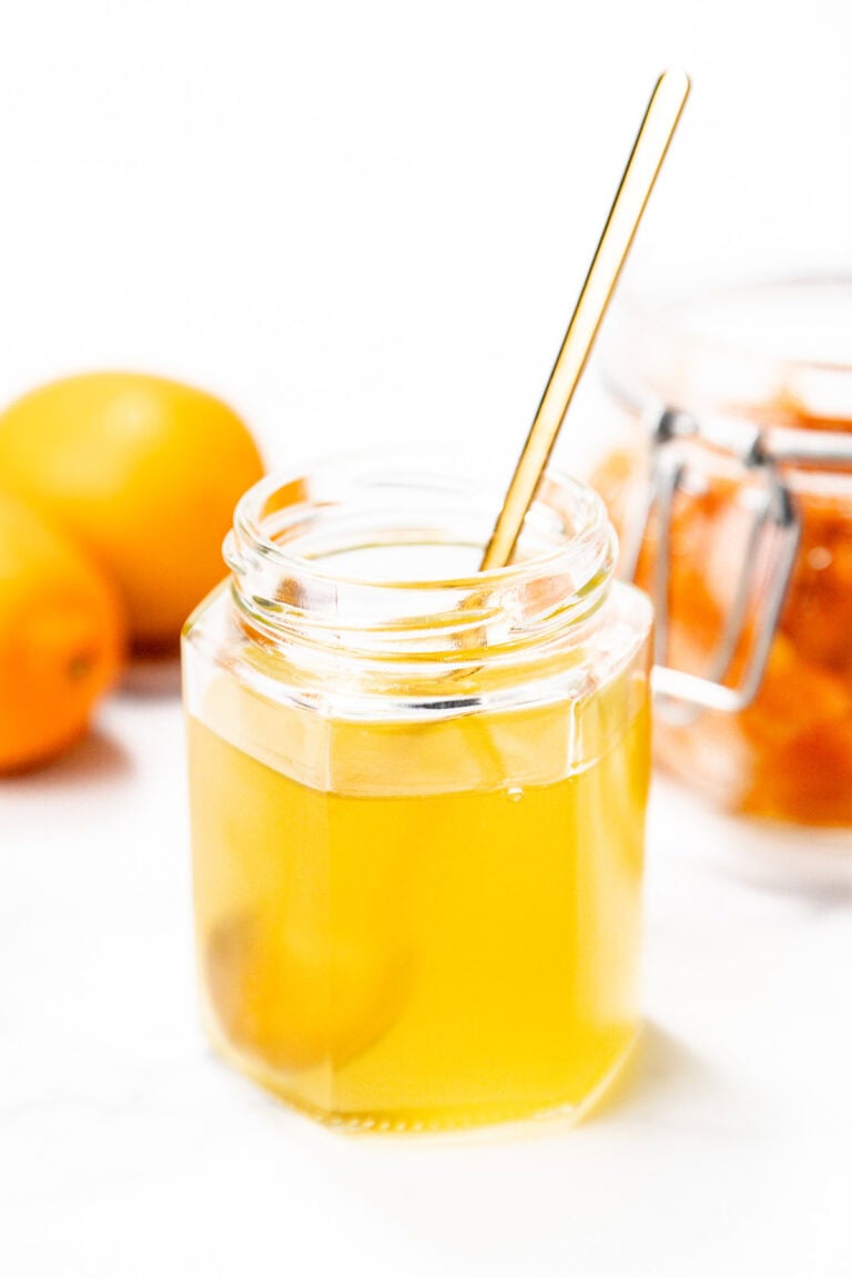 A hexagonal glass jar filled with golden honey, a metal spoon inside, with whole lemons and a jar of preserved lemons in the background.