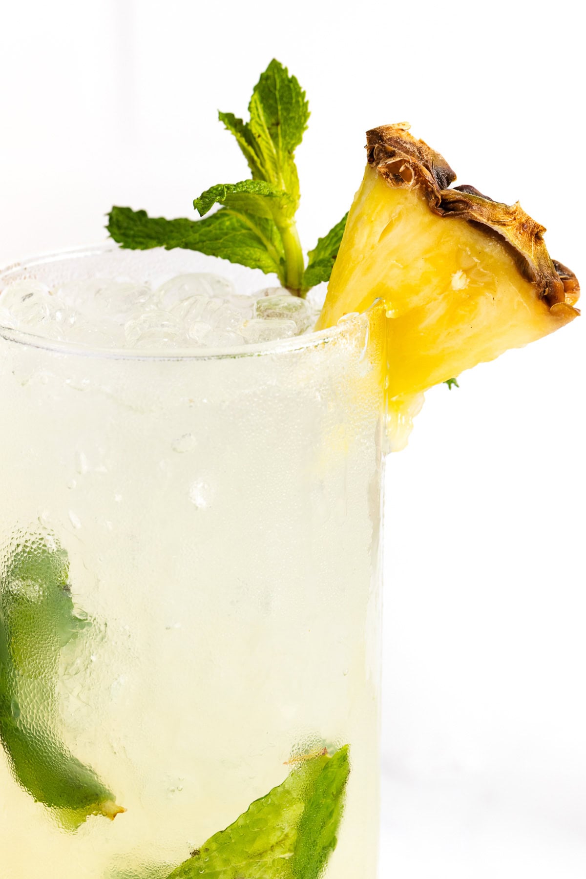 A cold drink with ice, mint leaves, and a pineapple wedge garnish in a clear glass, against a white background.