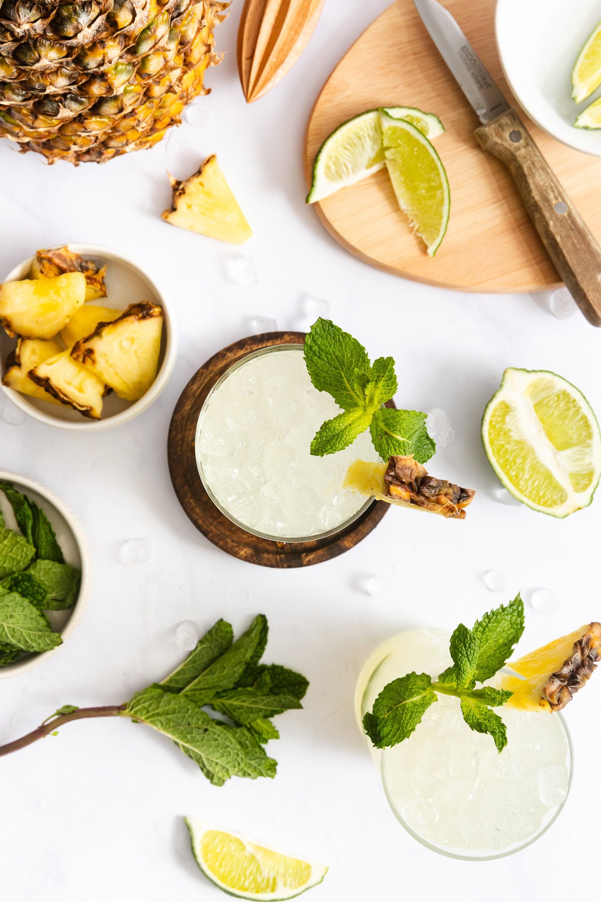 Two glasses of a cold drink garnished with mint and pineapple, surrounded by fresh lime wedges, pineapple pieces, mint leaves, and a pineapple top on a white surface.