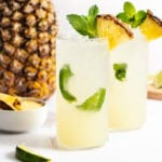 Two tall glasses of a yellow tropical drink garnished with mint, lime, and a pineapple wedge, with a whole pineapple and lime pieces in the background.