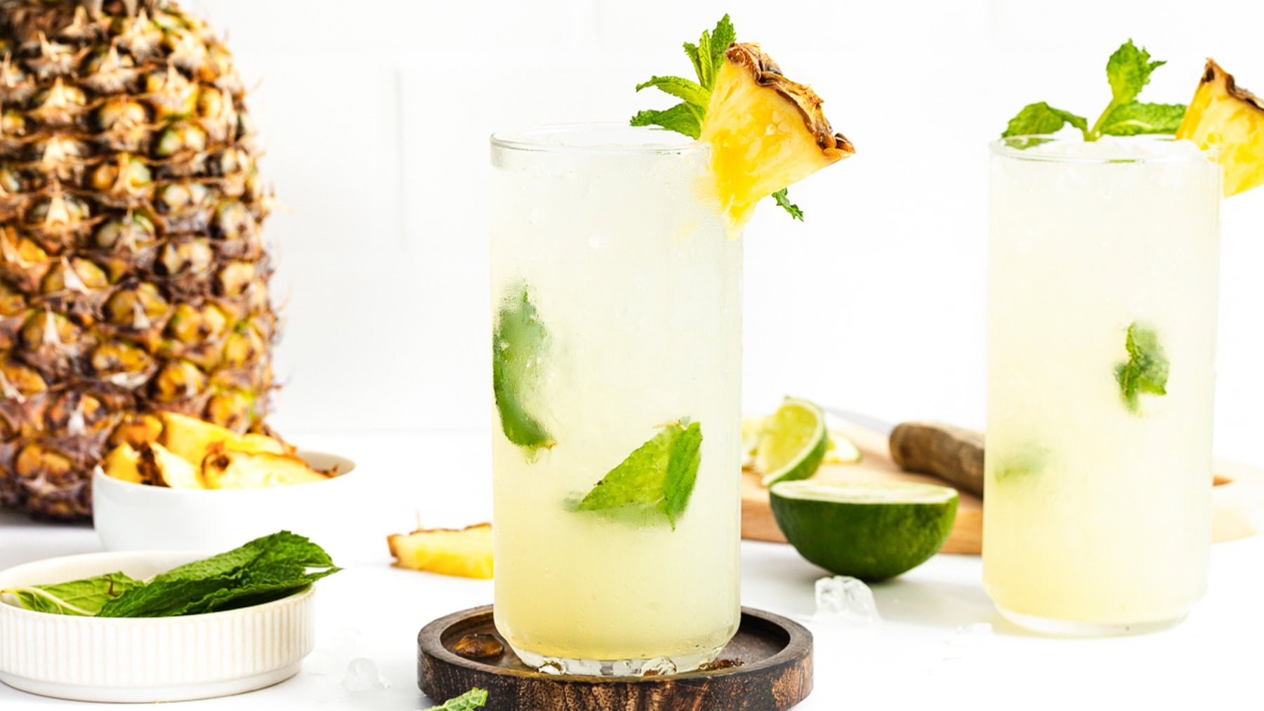 Two glasses of a light yellow drink garnished with mint and pineapple sit on a white surface, with fresh pineapple, lime, and mint leaves in the background.