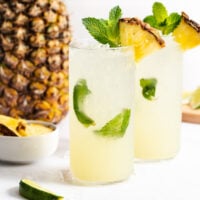 Two tall glasses of a yellow tropical drink garnished with mint, lime, and a pineapple wedge, with a whole pineapple and lime pieces in the background.
