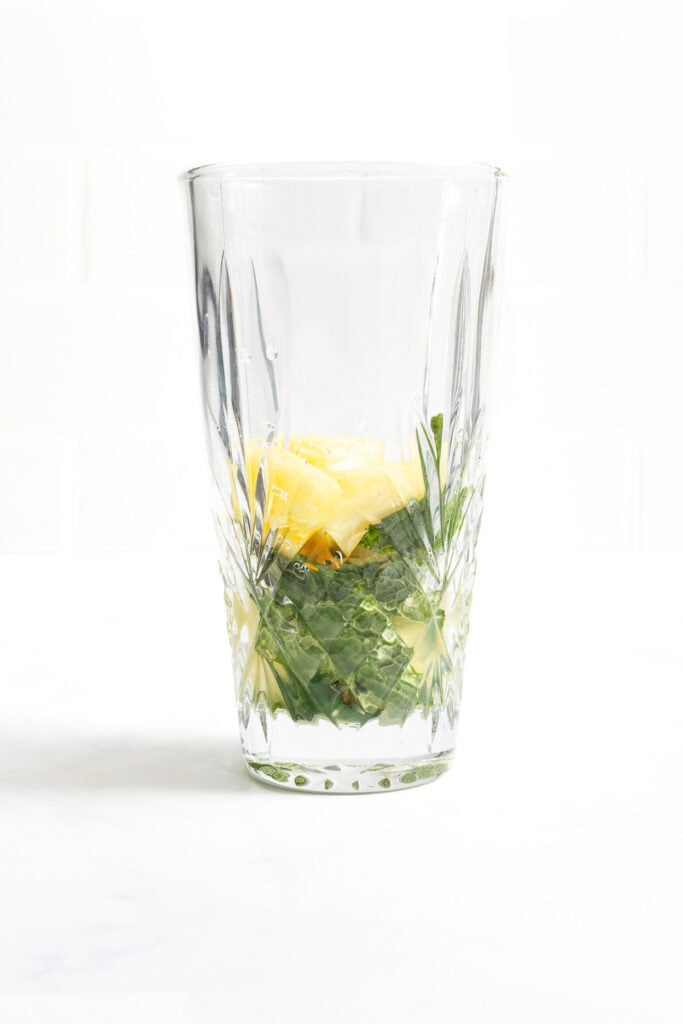 A clear glass containing chopped pineapple and mint leaves on a white surface with a white background.