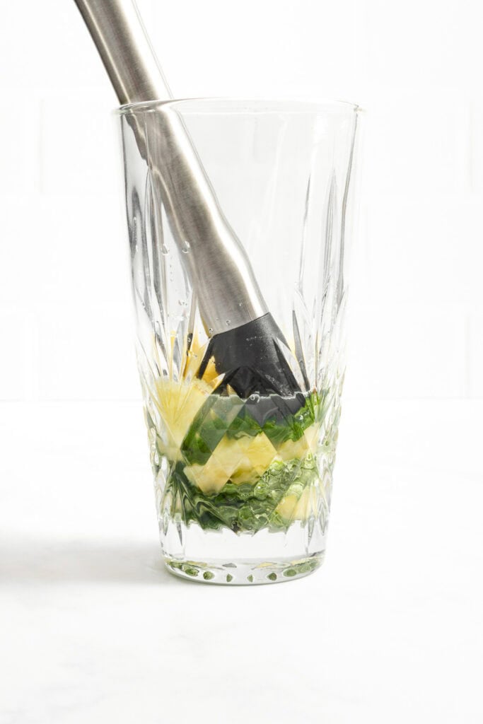 A metal muddler pressing pineapple chunks and mint leaves at the bottom of a clear, patterned glass.
