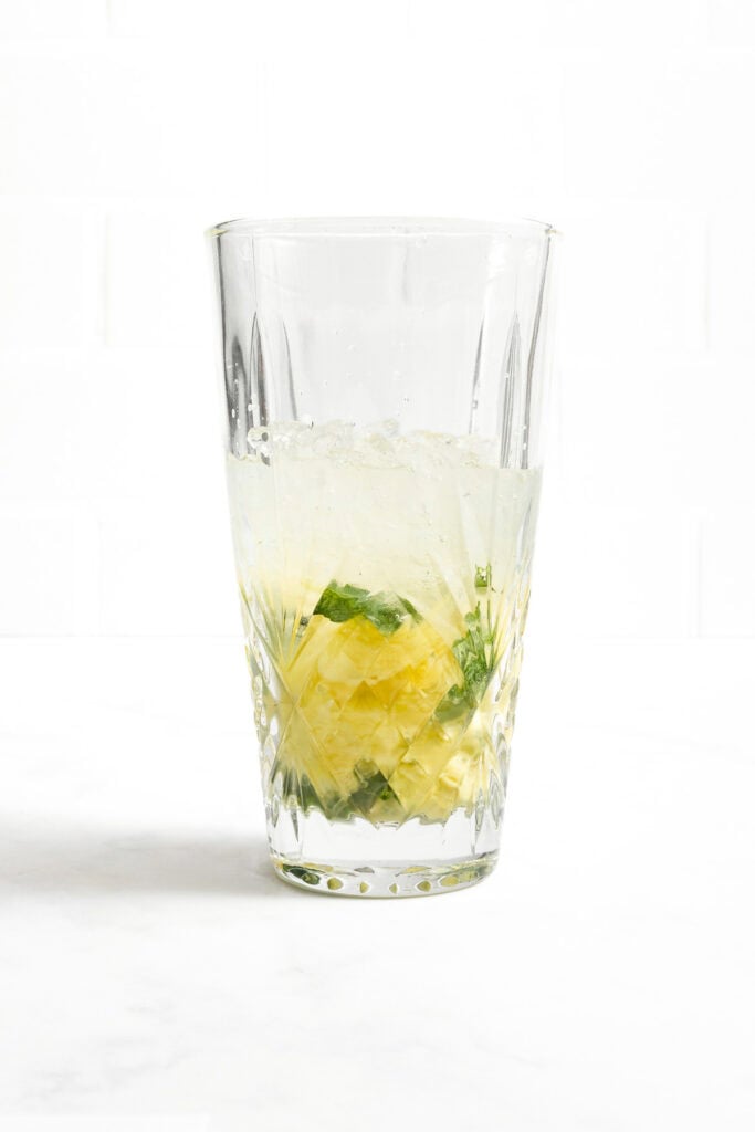 A clear glass with crushed ice, yellow fruit pieces, and green mint leaves on a white surface and background.