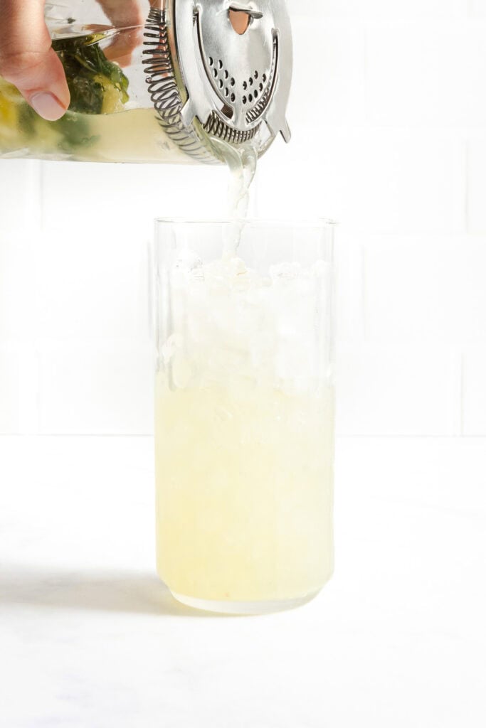A hand pours a yellow-green drink through a cocktail strainer into a glass filled with crushed ice against a white background.