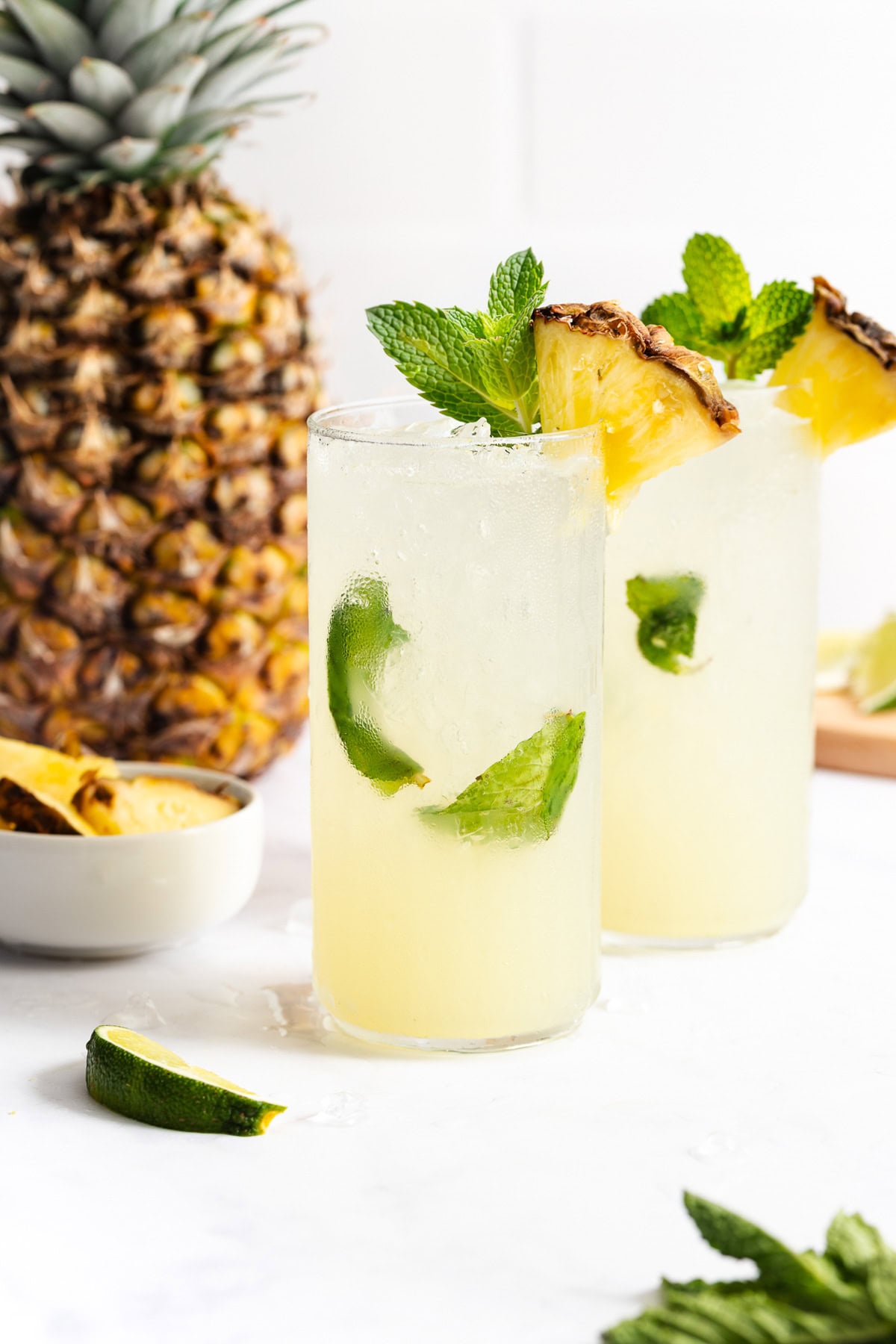 Two tall glasses of a yellow tropical drink garnished with mint, lime, and a pineapple wedge, with a whole pineapple and lime pieces in the background.
