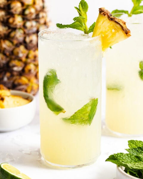 A glass of pineapple-lime cocktail with ice, garnished with a pineapple slice and mint; a pineapple and mint leaves are in the background.