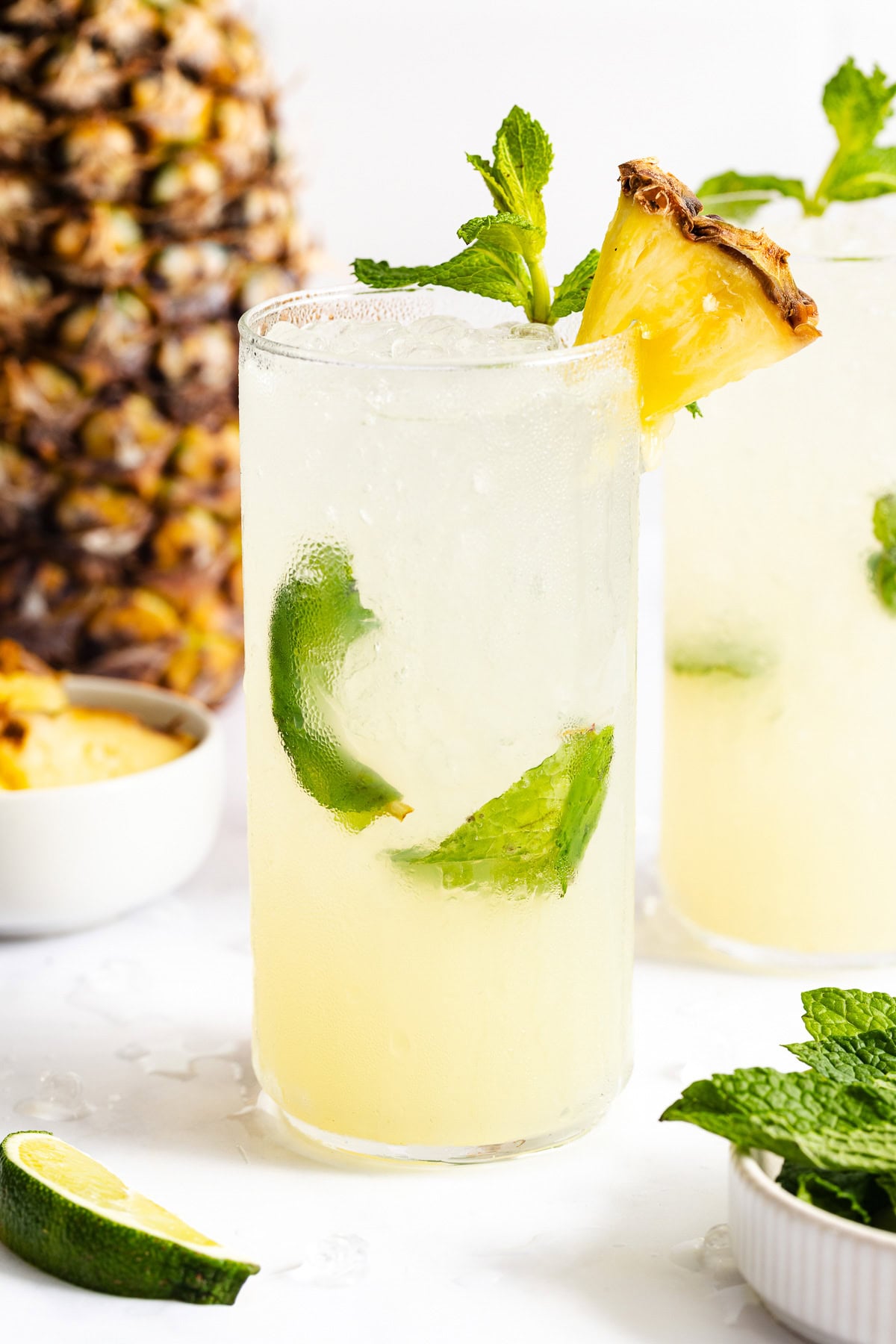 A glass of pineapple-lime cocktail with ice, garnished with a pineapple slice and mint; a pineapple and mint leaves are in the background.
