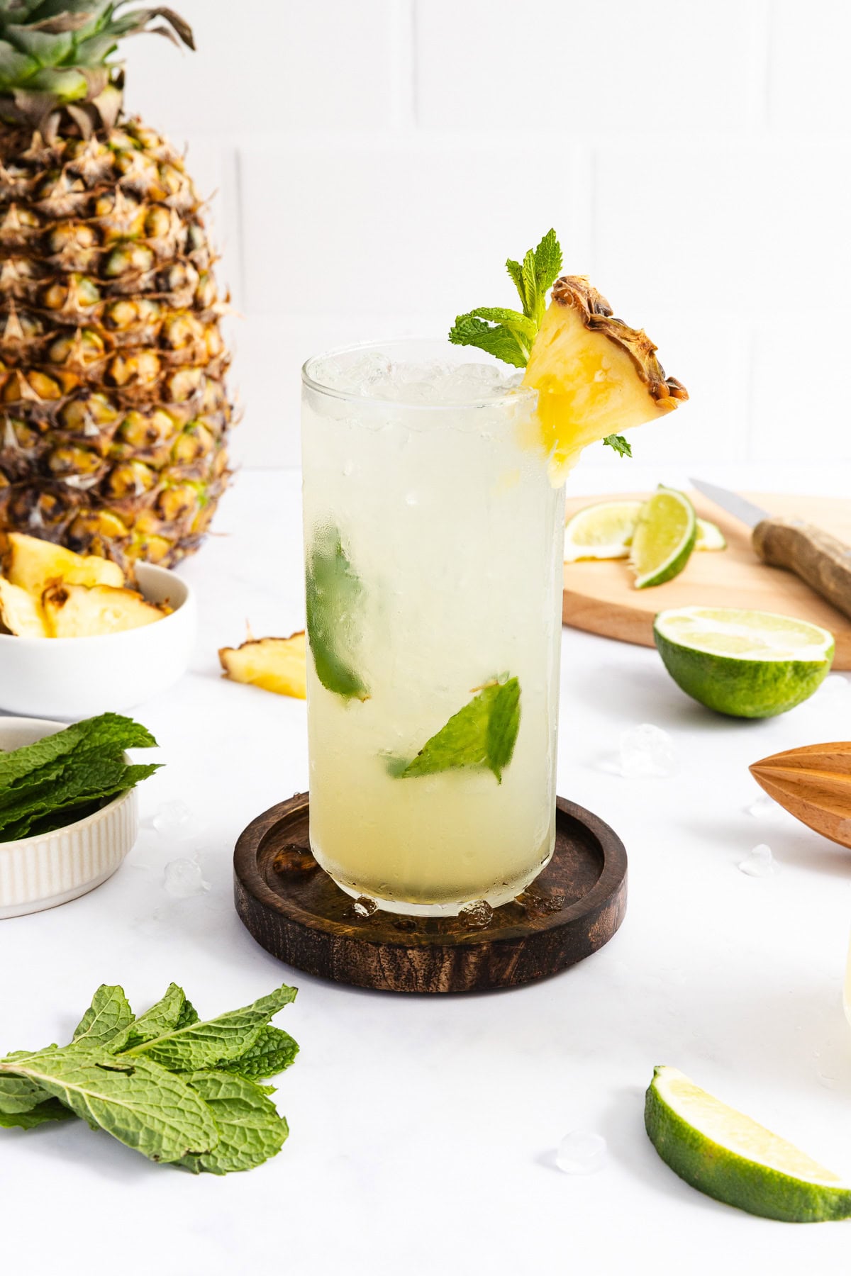 A tall glass of iced tropical drink garnished with a pineapple slice and mint, surrounded by limes, mint leaves, and a whole pineapple on a white surface.