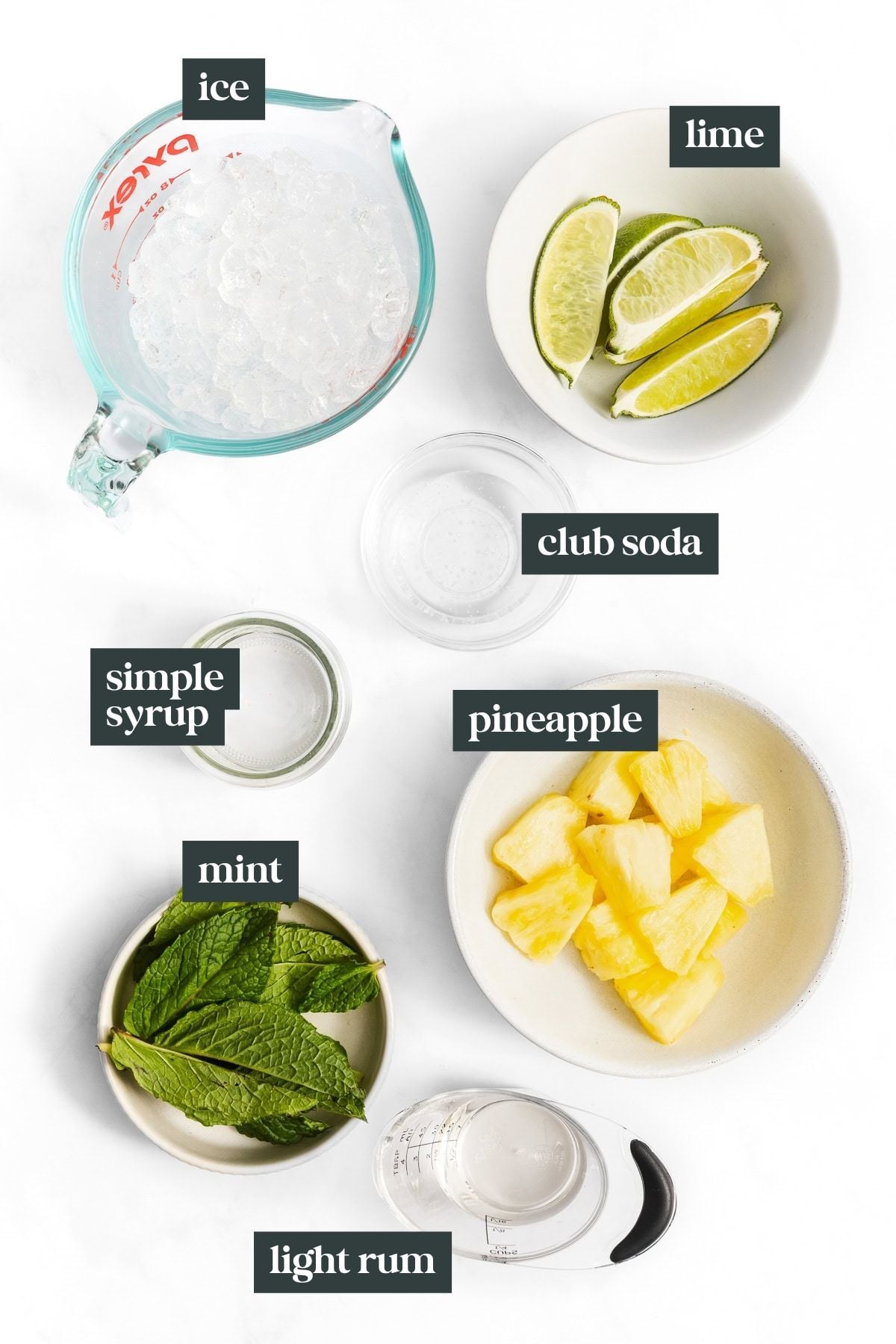 Overhead view of ingredients for a cocktail: ice, lime wedges, club soda, pineapple chunks, simple syrup, mint leaves, and light rum, each in separate bowls or containers.