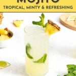 A glass of pineapple mojito garnished with mint and a pineapple wedge, surrounded by lime, pineapple pieces, and mint leaves on a white surface.