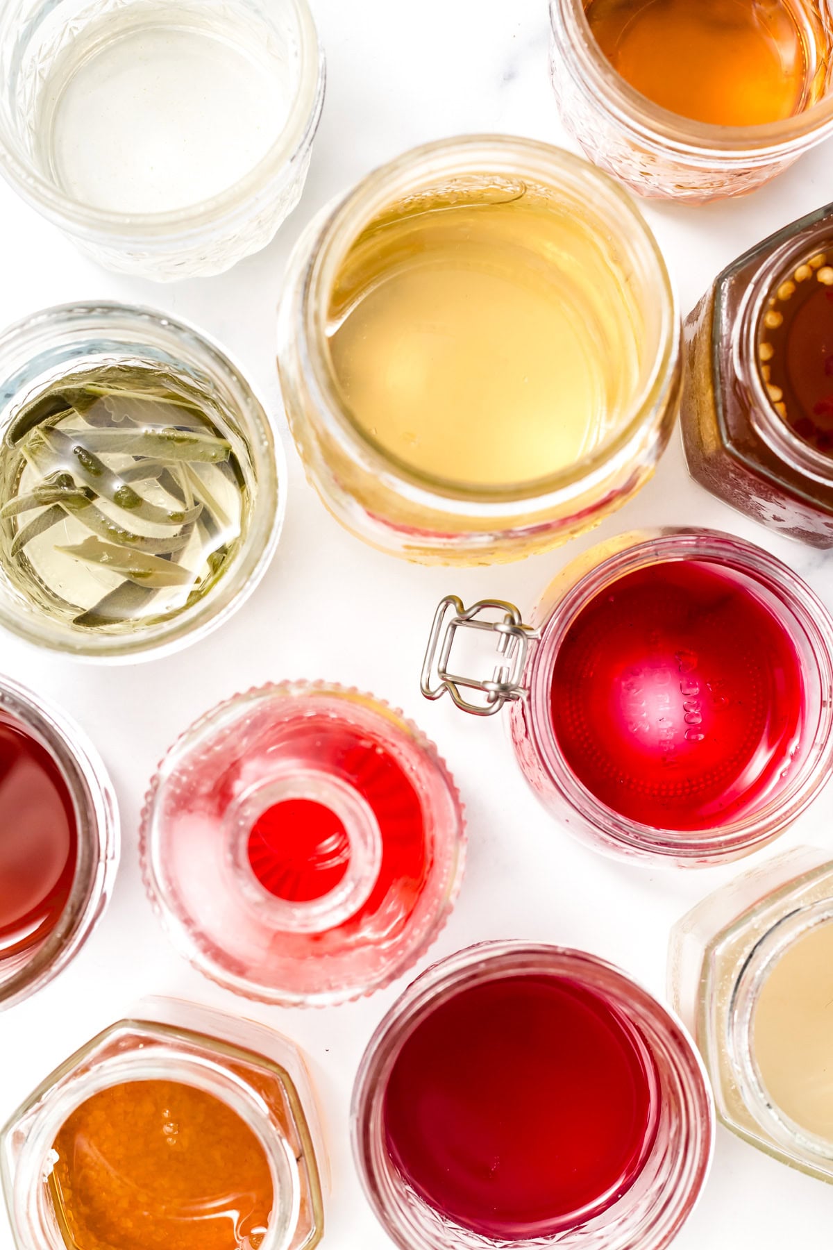 Assorted jars filled with colorful liquids and herbs, viewed from above on a white surface.