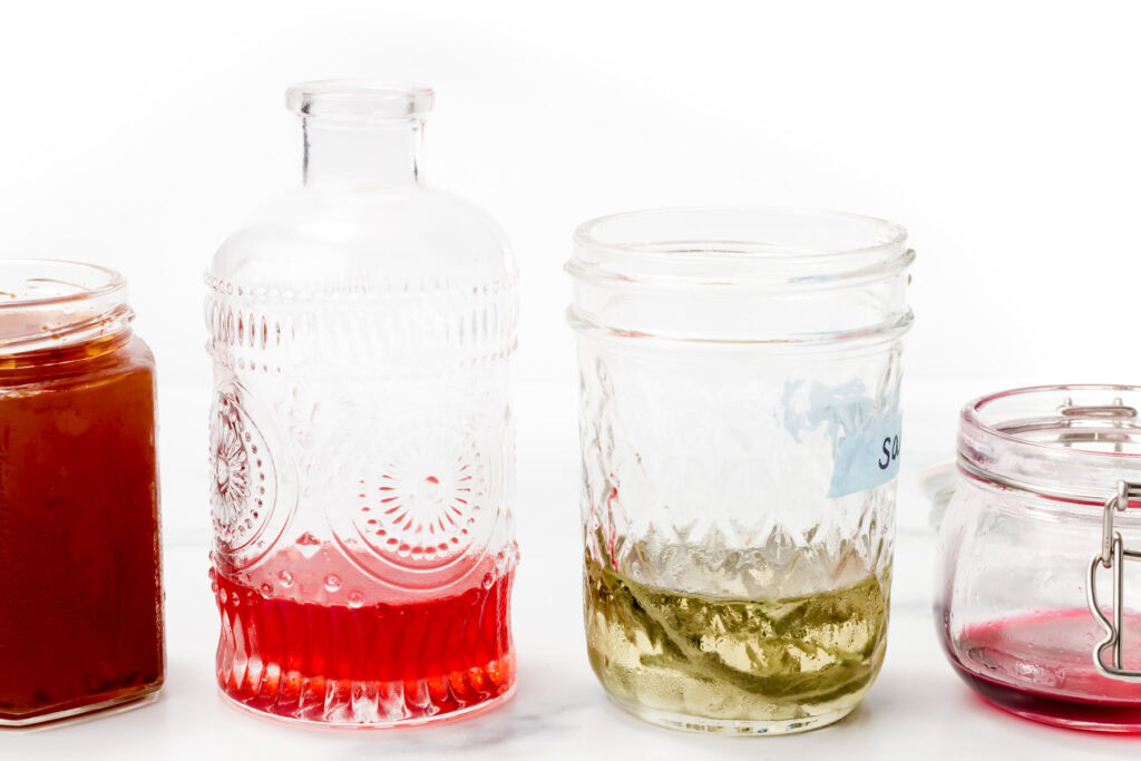 Four glass jars with colorful liquids, including red and yellow, arranged side by side on a white background.