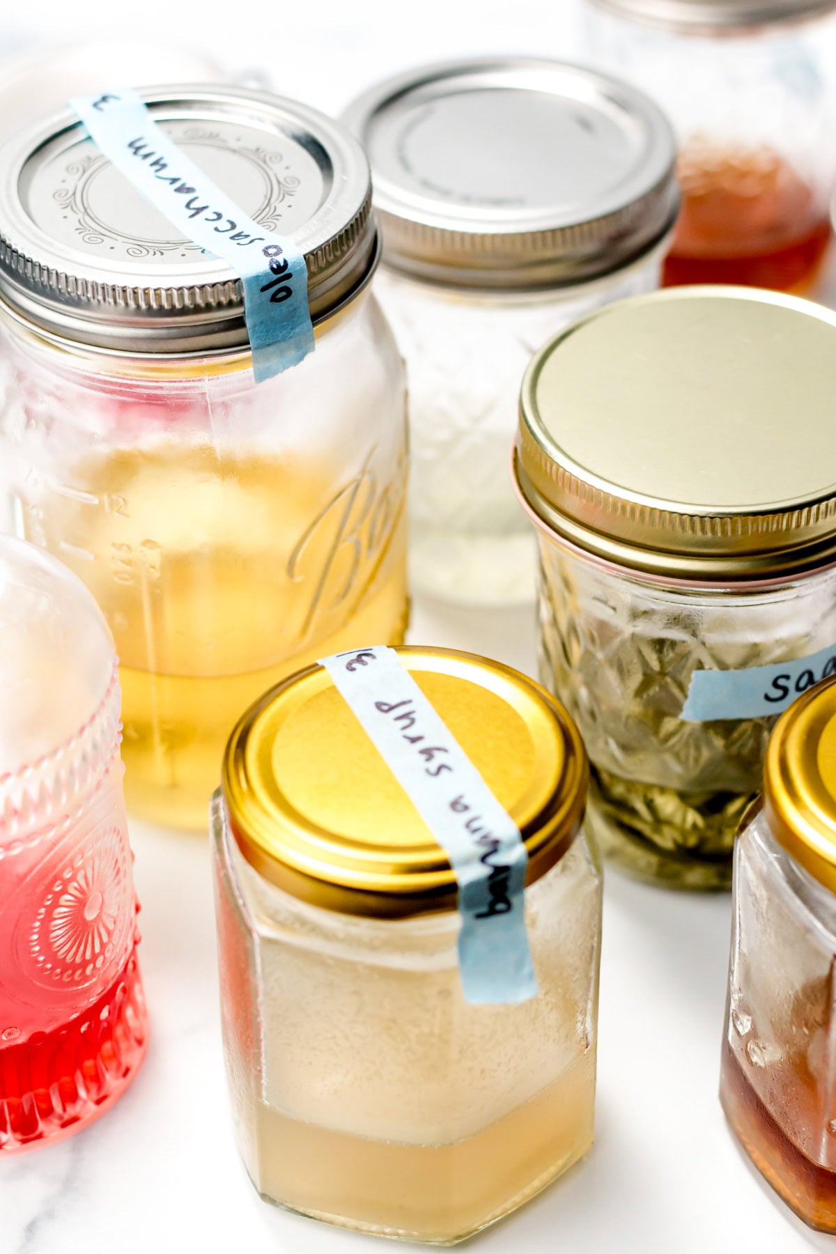 Assorted jars with metal lids and labels, containing homemade syrups or liquids on a white surface.