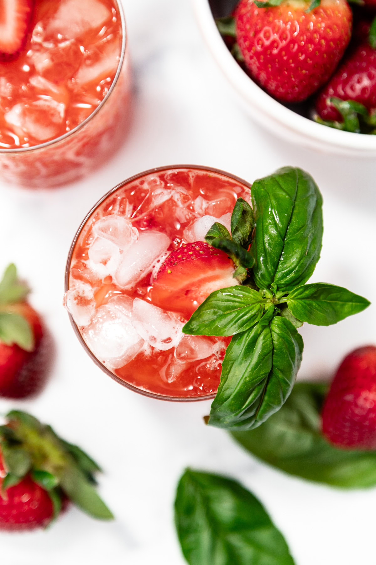 A glass of iced strawberry drink garnished with a fresh strawberry and basil leaves, surrounded by whole strawberries and basil on a white surface.